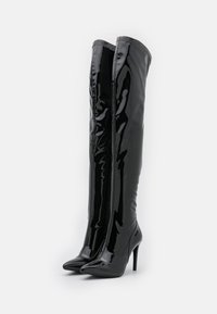 Botas de charol negro hasta el muslo con puntas afiladas y altos tacones de aguja, con una textura suave y brillante y cierre de cremallera.