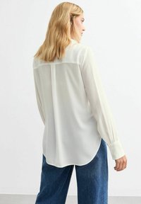 Blouse blanche à manches longues en tissu léger ; le dos présente un empiècement avec une couture centrale ; portée avec un jean bleu taille haute.