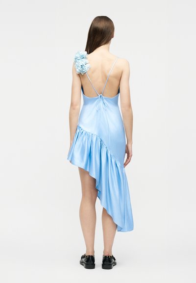 Γυναίκα που φοράει ανοιχτό γαλάζιο ασύμμετρο σατινέ φόρεμα με λεπτομέρεια λουλουδιών στον ώμο, λεπτές τιράντες, ανοιχτή πλάτη και μαύρα μοκασίνια, στέκεται με την πλάτη προς τα εμπρός.