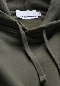 Olivgrüner Hoodie mit weicher Textur, verstellbaren Kordeln und einem Etikett mit der Aufschrift "ARMEDANGELS", das sich innen am Kragen befindet.