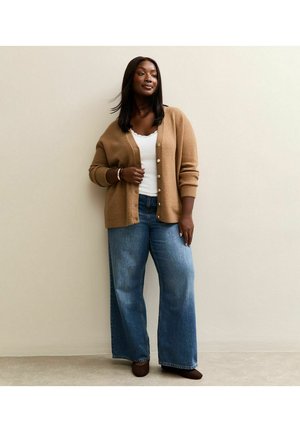 Donna che indossa un cardigan color sabbia, una maglia bianca, jeans blu a gamba larga e scarpe scure, in piedi contro un muro beige con un'espressione neutra.