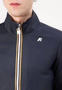 Homme portant une veste bleu marine avec un col montant, une fermeture éclair blanche encadrée de rayures jaunes et orange, et un logo géométrique blanc sur la poitrine.