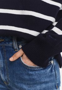 Tamno plavi prugasti pulover s bijelim horizontalnim prugama, rebrastim rukavima i pletenom teksturom, u kombinaciji s plavim trapericama.