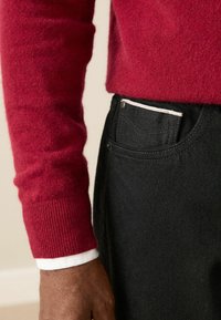 Red knit sweater with ribbed cuffs, worn over a white collared shirt, paired with black denim trousers featuring a white stitch detail on the pocket.
