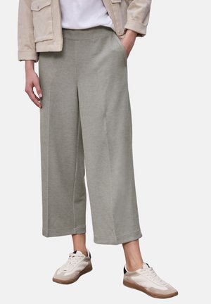 Person iført lysegrå, vide, cropped bukser, beige jakke, hvid skjorte og hvide sneakers med gummisåler, stående med den ene hånd i lommen.