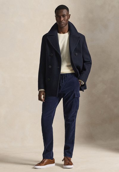 Man wearing navy pea coat over beige sweater, navy cargo pants, and brown leather sneakers, standing against plain neutral background.