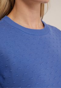 Blå stickad tröja med ett texturerat mönster av upphöjda prickar, rund halsringning och ribbad krage. Tyget ser slätt och mjukt ut.