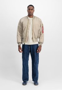 Beige bomber jacket with ribbed cuffs and hem, featuring a zip pocket and red tag. Paired with loose-fitting dark denim jeans.