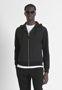 Giacca nera imbottita con zip e cappuccio, con maniche completamente nere e polsini a costine. Indossata sopra una maglietta bianca e abbinata a pantaloni neri.