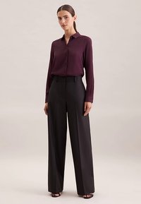 Bordeauxrode blouse met lange mouwen, een kraag en knoopsluiting, gecombineerd met zwart, hooggesneden broek. Gladde stof, getailleerde pasvorm, brede pijp.