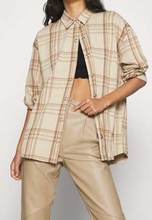 Kvinde iført beige ternet skjorte med knapper over sort crop top og beige læderbukser med høj talje, hånd hvilende på linningen.