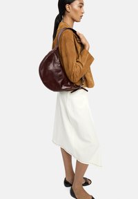 Ciemnoczerwony skórzany torebka w zaokrąglonym kształcie, z paskiem na ramię i strukturalnym wykończeniem, zestawiona z jasnobeżową spódnicą i brązową kurtką.