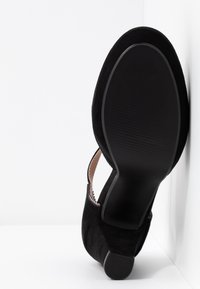 Chaussure de ville femme en daim noir avec talon bloc, semelle tournée vers l'avant, positionnée sur un fond blanc en coin.