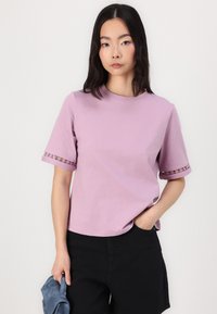 Lavendel bomull T-skjorte med korte ermer som har en blondedetalj ved ermene, sammen med svarte shorts og holder et blått tilbehør.