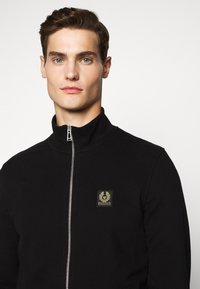 Svart zip-up jacka med hög krage, med en fyrkantig guld- och svart logotäppa på vänster sida och en slät tygstruktur.