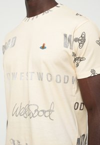 Tricou bej cu text gri "Westwood" imprimat de mai multe ori, logo mic în formă de orb colorat pe piept și modele negre de orb repetate pe mânecă.