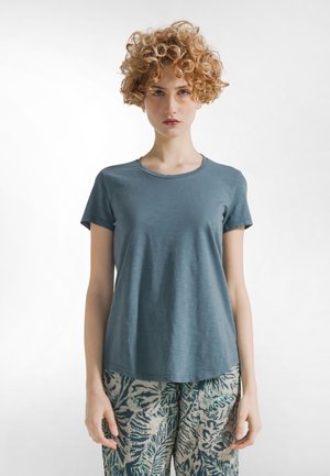 Tricou bleu deschis cu mâneci scurte, realizat din material moale, cu croială lejeră și decolteu rotund. Asortat cu pantaloni largi, cu imprimeu.