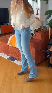 Femme aux cheveux longs portant un pull crème et un jean bleu, debout à l'intérieur près d'un canapé orange et un sol en bois.