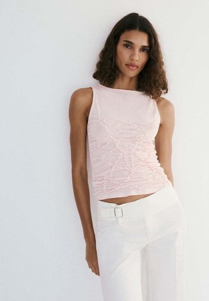Femme aux cheveux bouclés portant un haut sans manches rose clair avec un design texturé et un pantalon blanc taille haute avec ceinture, debout contre un mur blanc.