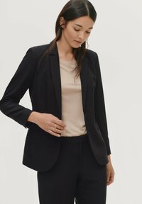 Zwarte op maat gemaakte blazer met notch-kleppen en twee knopen, gecombineerd met een beige top. Beschikt over een gestroomlijnd ontwerp en een soepele stof.