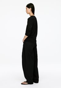 Schwarzes Langarmshirt mit hochgerollten Ärmeln, kombiniert mit lockeren schwarzen Hosen. Das Model steht im Profil und trägt schwarze Flip-Flops.