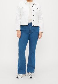 Person som bär en vit skjortjacka, beige topp, blå vida jeans och vita sneakers som står mot en enfärgad vit bakgrund.