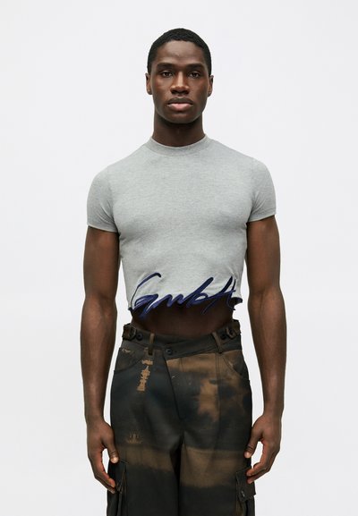 Vyras dėvi prigludusią pilką trumpą marškinių versiją su tamsiai mėlynu tekstu ir aukštai juosmeniui pritaikytas tamsias kroso kelnes su rusvaisiais pleistrais.