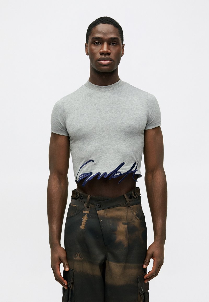 Man in een grijze, strakke cropped t-shirt met marineblauwe tekst en hooggesneden donkere cargo-broek met bruinachtige vlekken.