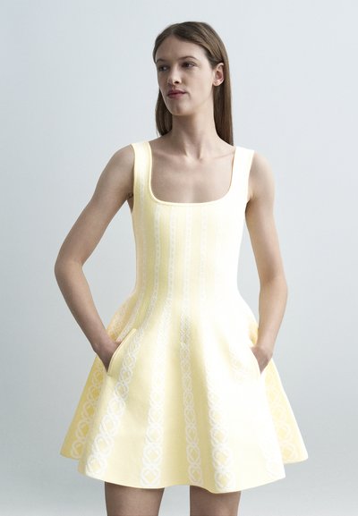 Femme portant une robe trapèze sans manches de couleur jaune pâle avec des motifs blancs verticaux subtils, debout les mains dans les poches devant un fond uni.