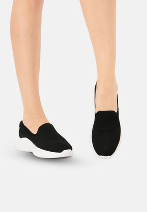 Voeten met zwarte slip-on sneakers met witte zolen, staand op een lichtgrijze ondergrond.