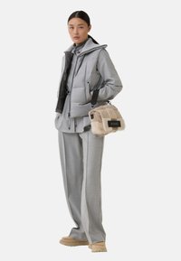 Frau steht in einem hellgrauen, schichtweise gestuften Outfit mit gesteppter Weste und weitem Hosen, hält eine beige und tanfarbene Schaffellhandtasche mit schwarzem Riemen.