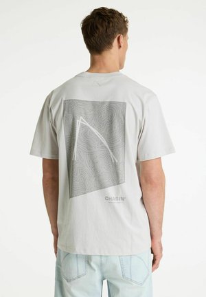 Homme portant un T-shirt gris clair avec un motif topographique abstrait et le logo "CHASIN" au dos, associé à un jean en denim bleu clair.