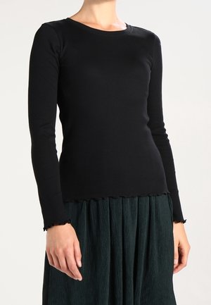 Femme portant un haut noir moulant à manches longues avec une finition en bordure en forme de laitue, associée à une jupe plissée vert foncé.