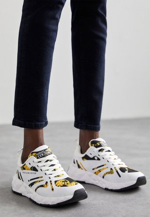 Personne portant un pantalon foncé et des baskets blanches Versace Jeans Couture avec des motifs baroques noirs et dorés, debout sur un sol gris.
