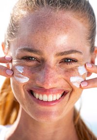 Donna con pelle chiara e lentiggini che applica una crema bianca sul viso con le dita, sorridendo, con occhi azzurri e capelli castani raccolti in una coda di cavallo.