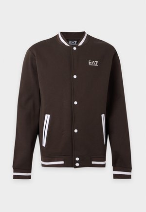 Homme portant une veste noire EA7, un polo noir, un pantalon beige, des baskets beiges et une casquette blanche EA7, debout devant un fond uni.