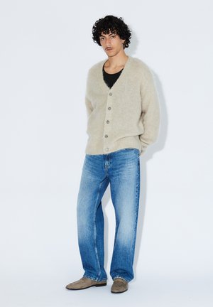 Beige, blød, strikket cardigan med V-hals og knaplukning fortil, båret over en sort tanktop. Lyseblå, afslappede jeans og beige sko.