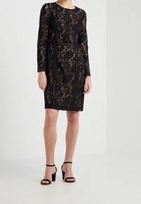 Kvinna iklädd en knälång, långärmad svart spetstklänning med blommönster och svarta högklackade sandaler, stående mot en vit vägg.