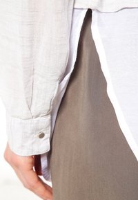 Camicia bianca a maniche lunghe con tessuto testurizzato, dotata di polsino con bottoni e sovrapposta a un tessuto grigio liscio nella parte inferiore.