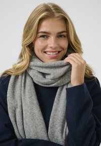 Bufanda de lana suave y gris drapeada alrededor del cuello, con una textura suave. El modelo lleva un abrigo azul marino, con maquillaje natural y cabello rubio.