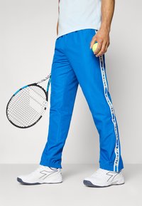 Blauwe sportbroek van lichtgewicht materiaal met elastische boorden en zijstrepen met de tekst "LACOSTE." Witte sneakers met rubberen zool.