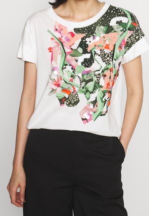 Hvid kortærmet top med pink, grøn og sort blomster- og abstrakt lap-design, båret med sorte bukser, den ene hånd i lommen.