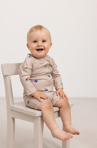 Beige babysvømmedrakt med lange ermer, med blå og lyseblå mønstre, rund halsutringning og korte ben. Sitter på en lys stol.
