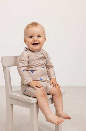 Maillot de bain beige pour bébé avec manches longues, présentant des motifs bleus et bleu clair, un col rond et des jambes courtes. Assis sur une chaise claire.
