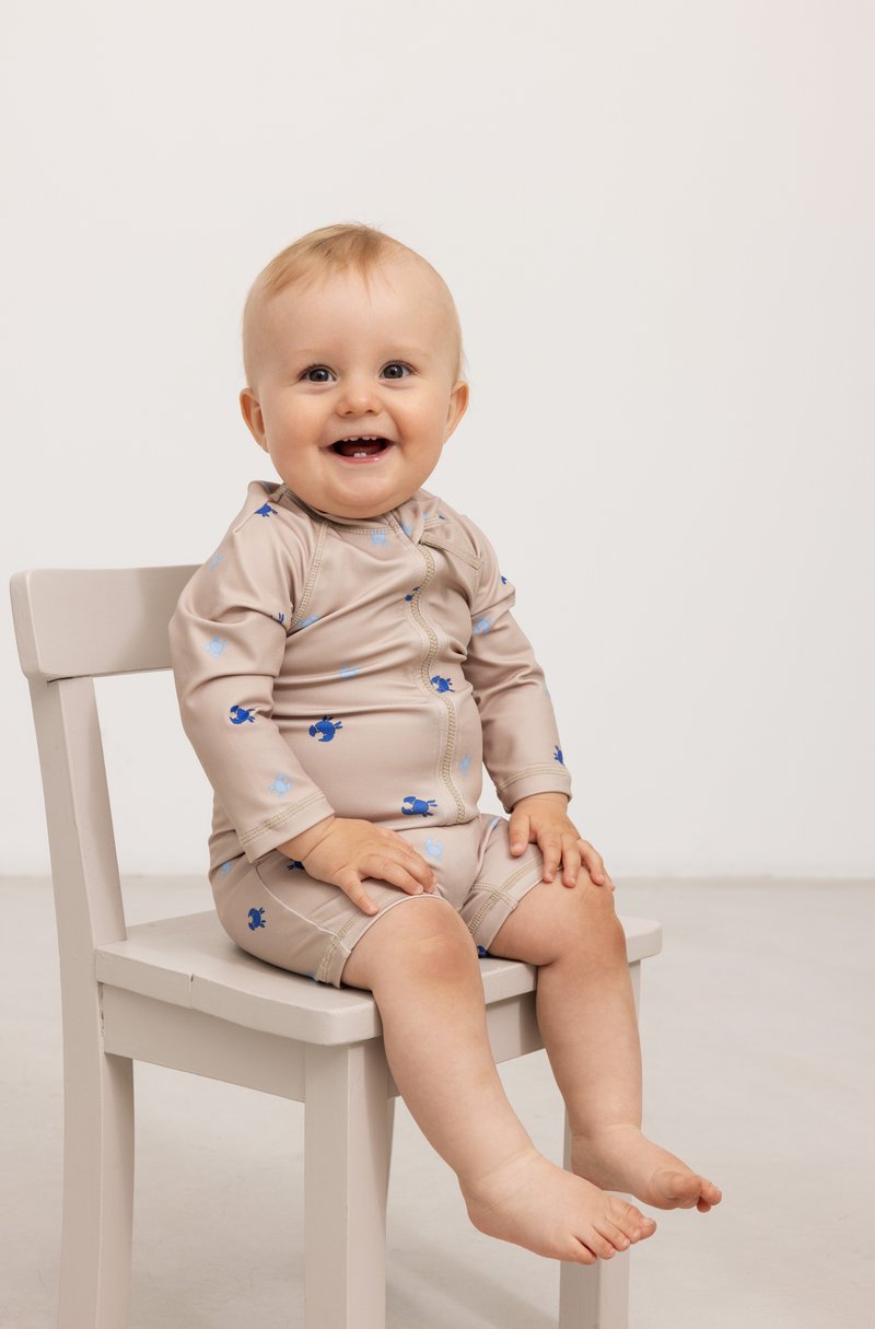 Beige babysvømmedrakt med lange ermer, med blå og lyseblå mønstre, rund halsutringning og korte ben. Sitter på en lys stol.