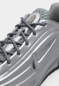 Κοντινό πλάνο μεταλλικού ασημί αθλητικού παπουτσιού με γκρι κορδόνια, υφασμάτινη πράσινη γλώσσα με υφή και ανάγλυφο λογότυπο Nike στη πλευρά.