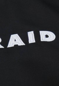 Schwarzer Stoff mit weißem gesticktem Text "RAID". Die Textur ist glatt, mit mattem Finish, und die Buchstaben sind leicht erhaben.