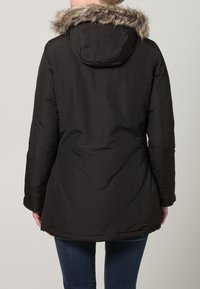 Černá parka s kapucí lemovanou umělou kožešinou, se rovný střihem, dlouhými rukávy a lehce zakřiveným spodním lemem. Hladký, voděodolný materiál.