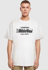White cotton t-shirt with a round neck, oversized fit, featuring black "COMPTON Athletics" logo centered on the chest, smooth texture.