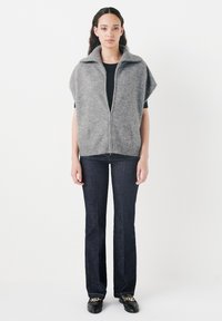 Grå, zippered väst med bred krage, bärs över en svart topp, parat med mörka denimjeans och svarta loafers med en guldkedjeaccent.